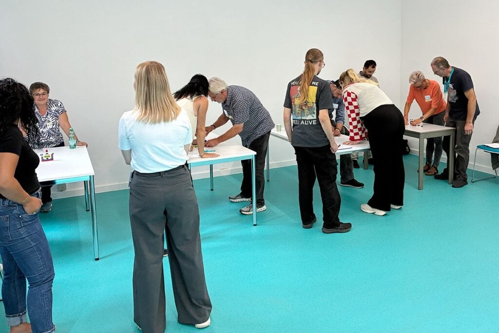 Mehrere Personen stehen in Gruppen an kleinen Tischen in einem Seminarraum und lösen gemeinsam Aufgaben